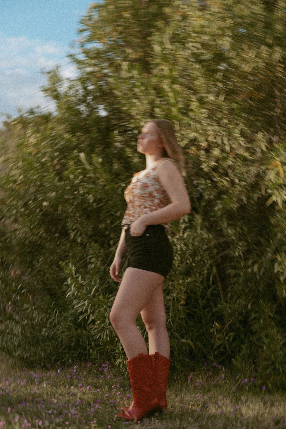 Person in brown cowboy boots and shorts stands against green foliage background in warm summer light.