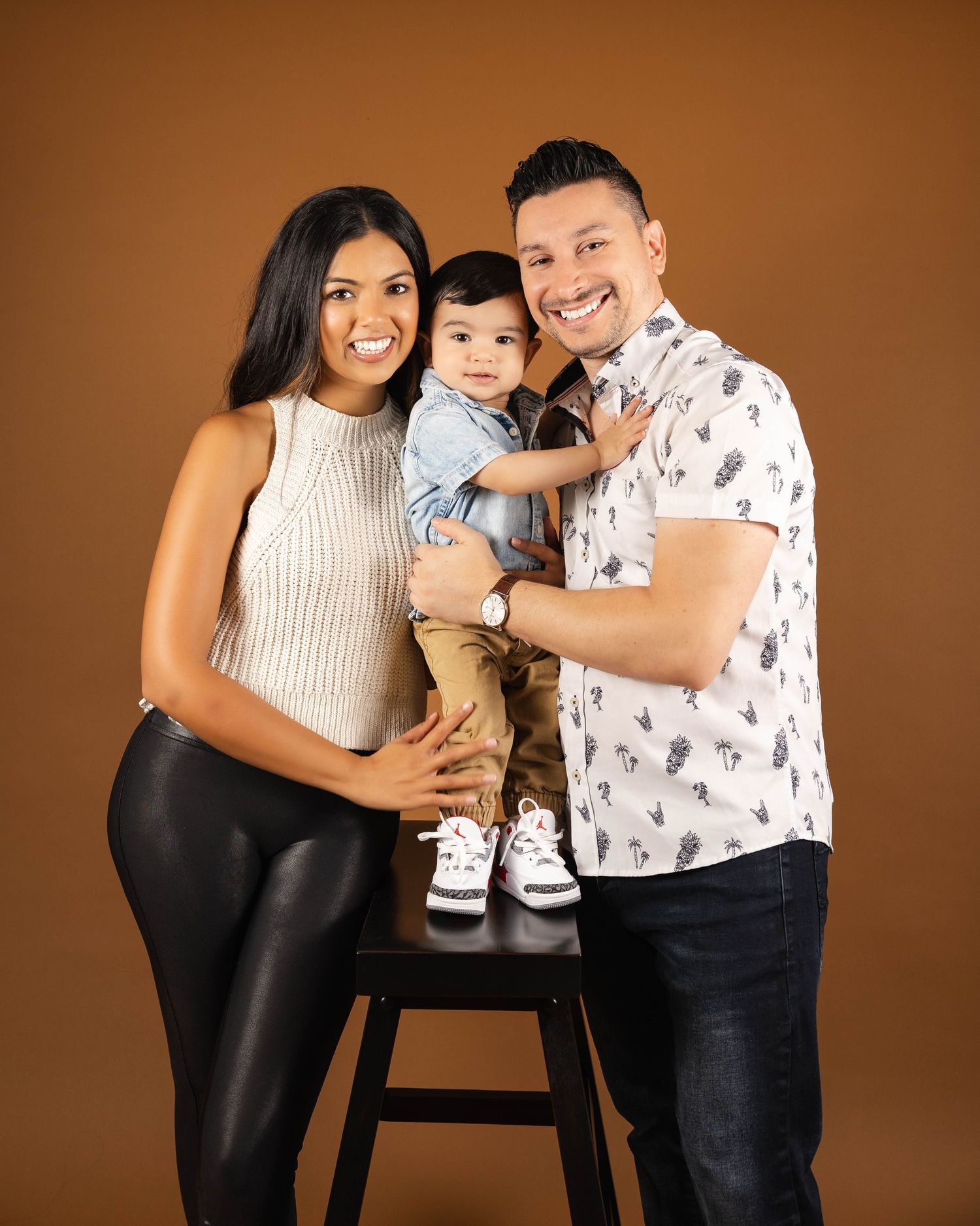 A happy family posing together against a warm brown backdrop, with a small child standing on a black stool between them.