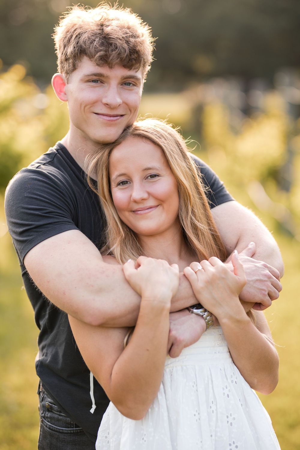 A couple embraces outdoors in golden sunlight, with her wearing a white dress and him in a black t-shirt.