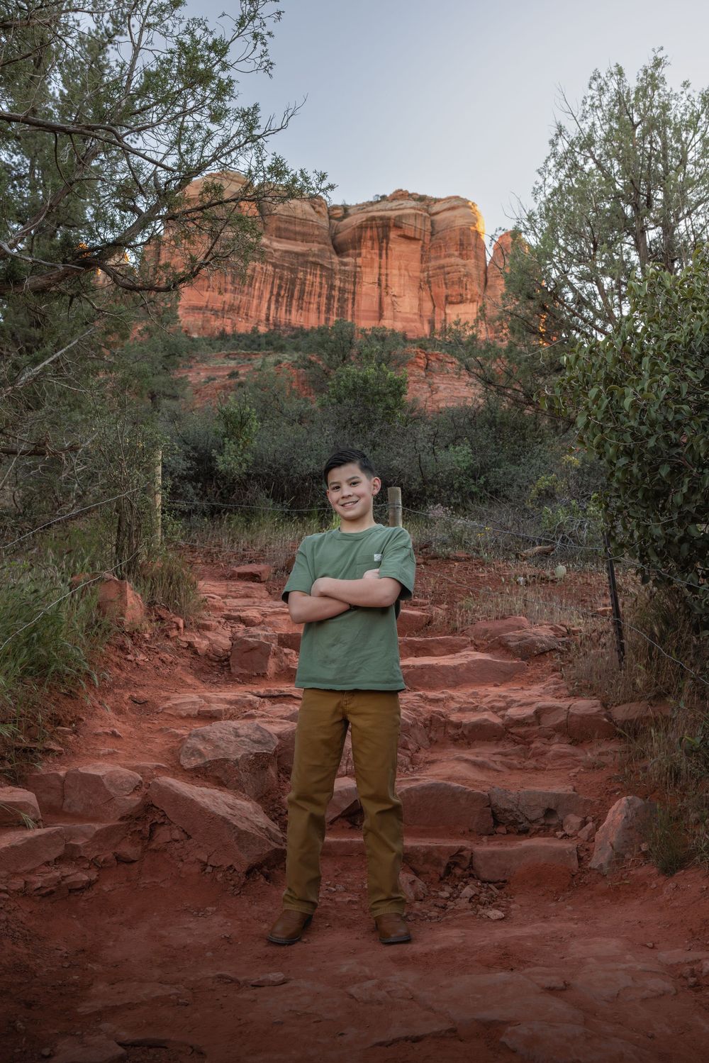 A smiling boy with arms crossed stands on red rock steps with Sedona's iconic red rock formations behind him.