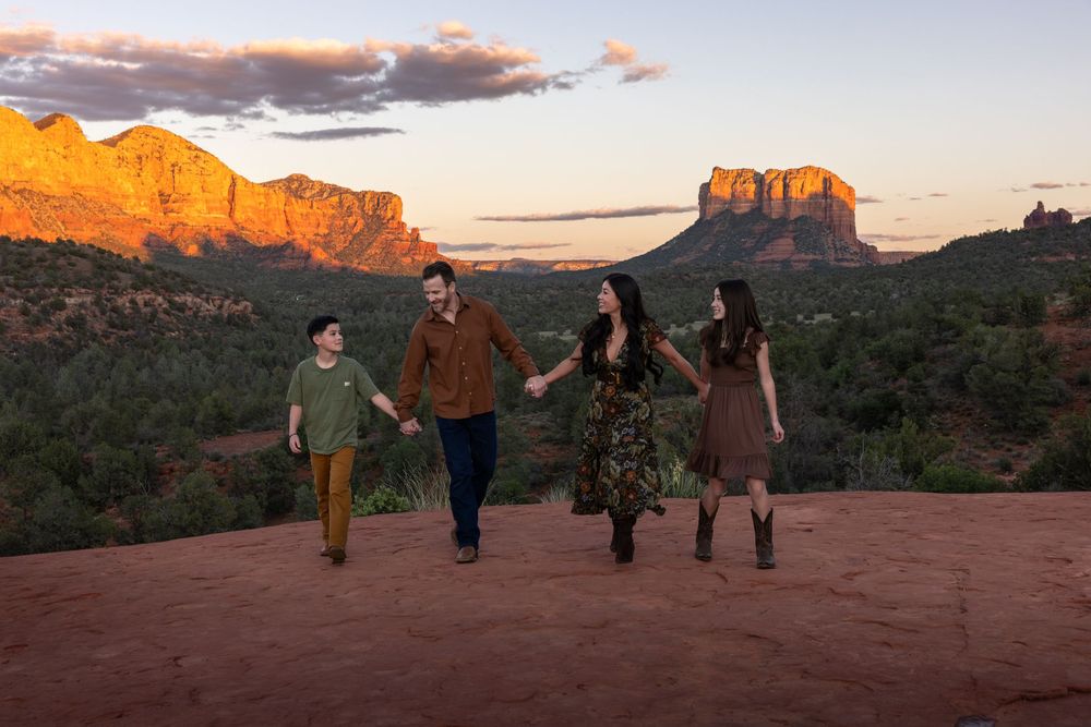 Family of four holding hands walking across red rock with glowing Sedona mountains at sunset.