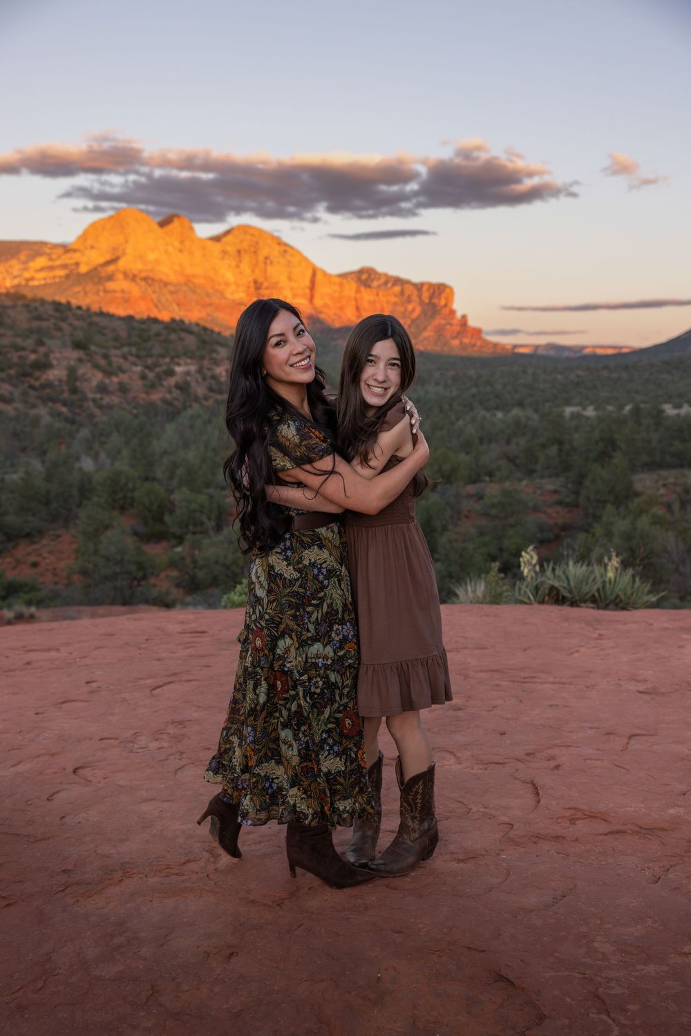 Mother and daughter hugging and smiling with golden Sedona red rock mountains at sunset behind them.