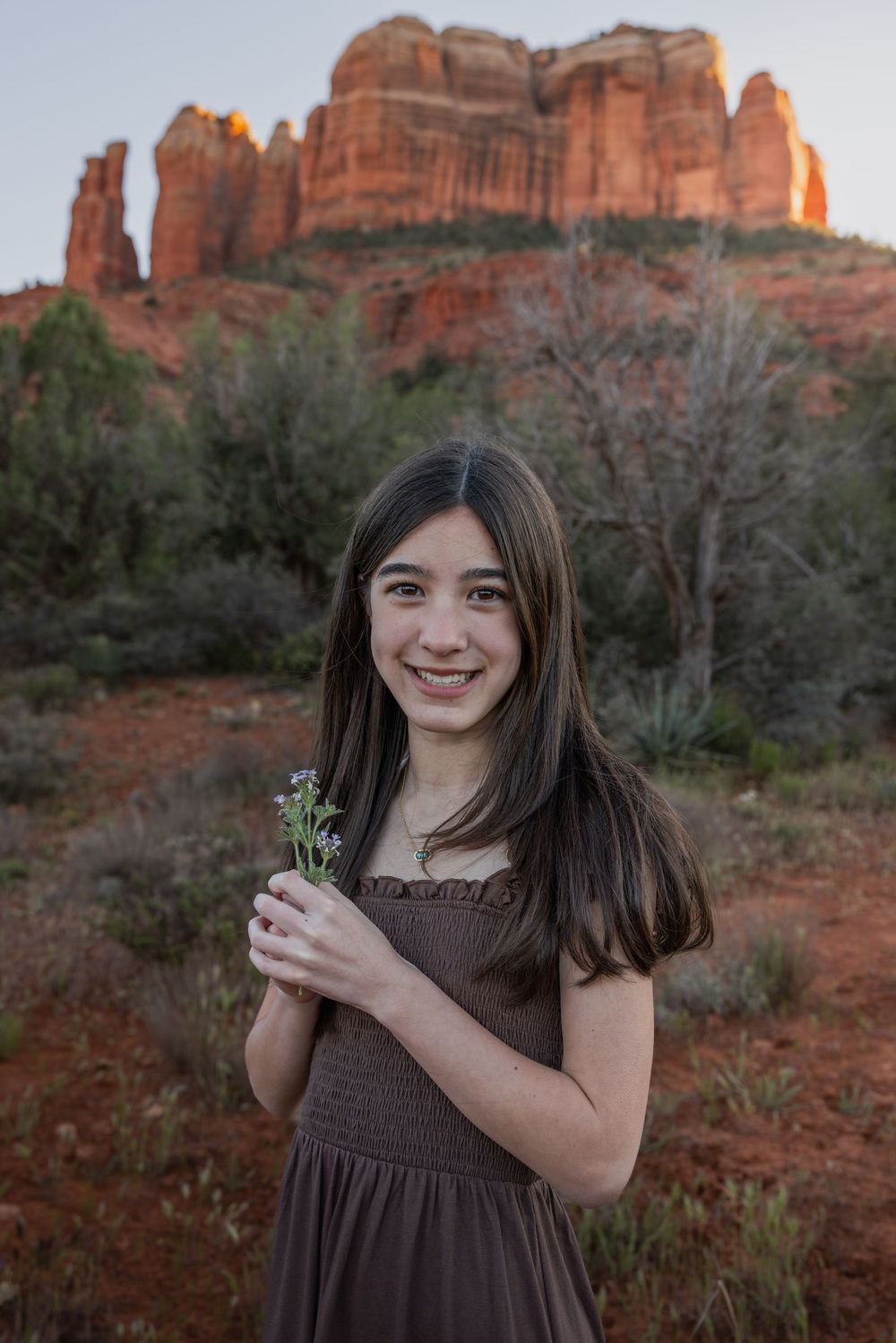 Young girl in brown dress holding wildflowers with Cathedral Rock in Sedona glowing at sunset.