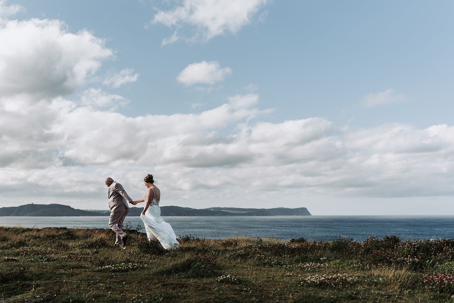 A couple walks along a coastal cliff with dramatic ocean views and puffy clouds in the blue sky.