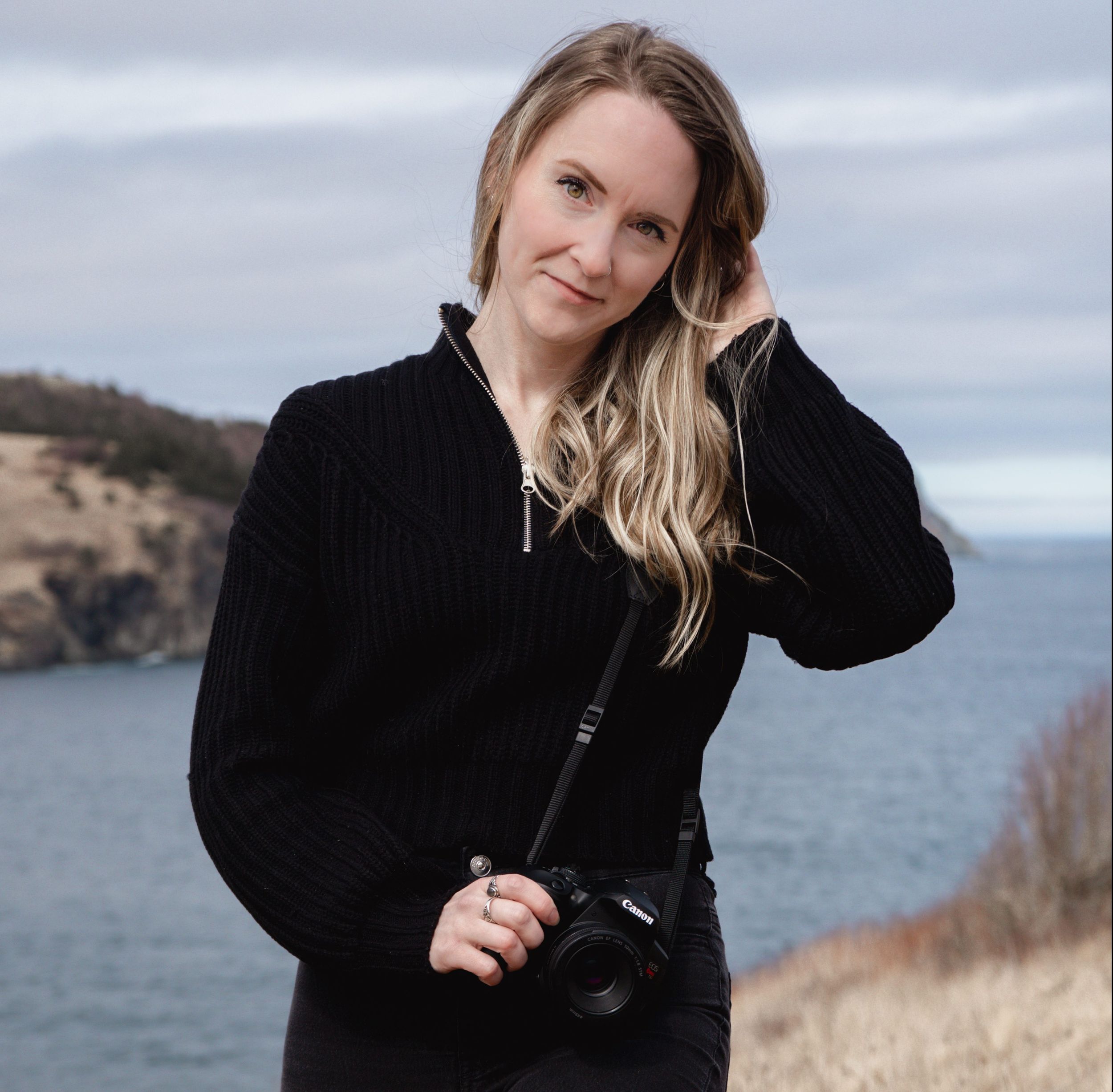 Person wearing a black sweater and carrying a camera stands on coastal cliffs overlooking the ocean on an overcast day.