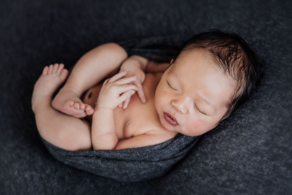 Peaceful sleeping newborn baby curled up on dark gray textile background during professional photoshoot.