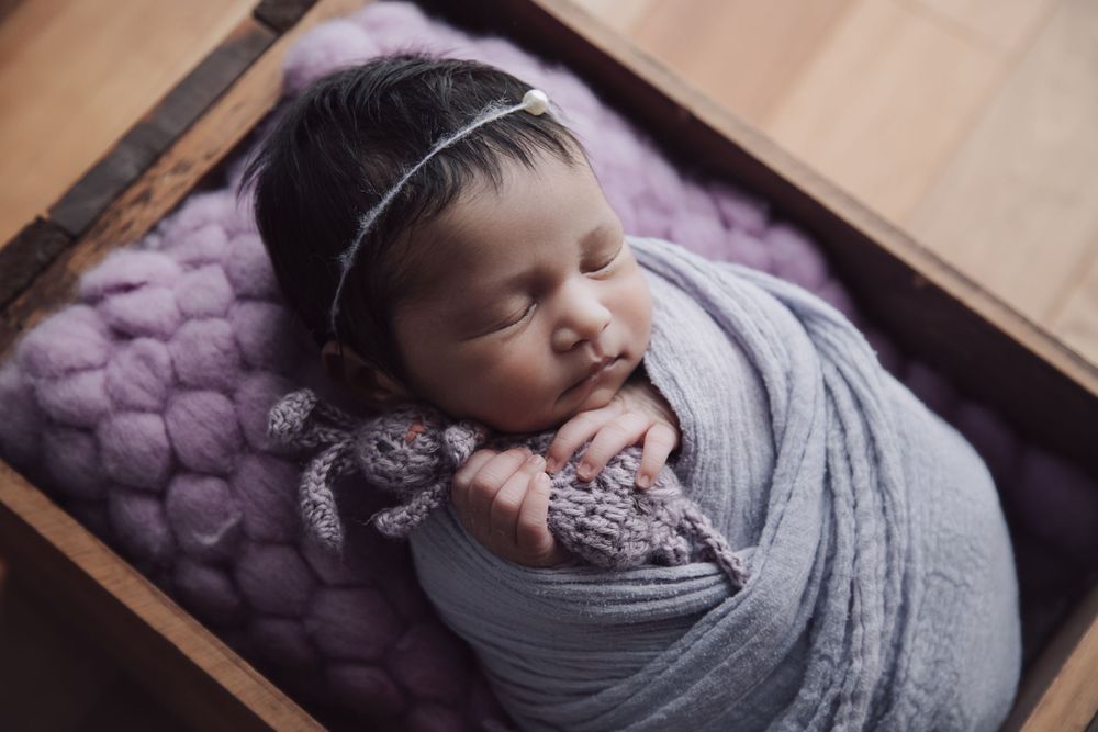 Newborn baby sleeping peacefully on purple blanket wrapped in soft grey swaddle.