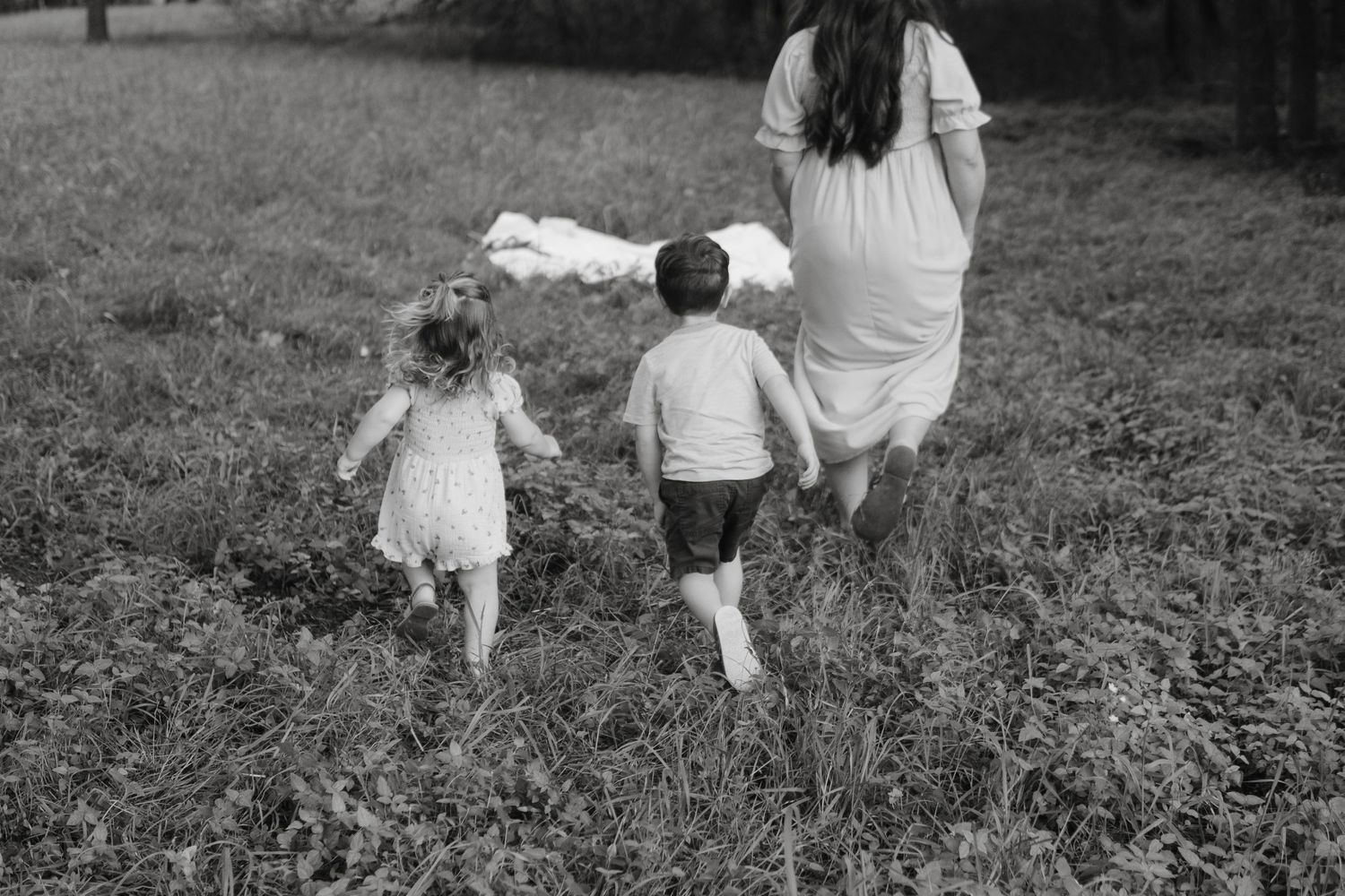A black and white photo shows two children following their mother while running through a grassy field together in a carefree manner.