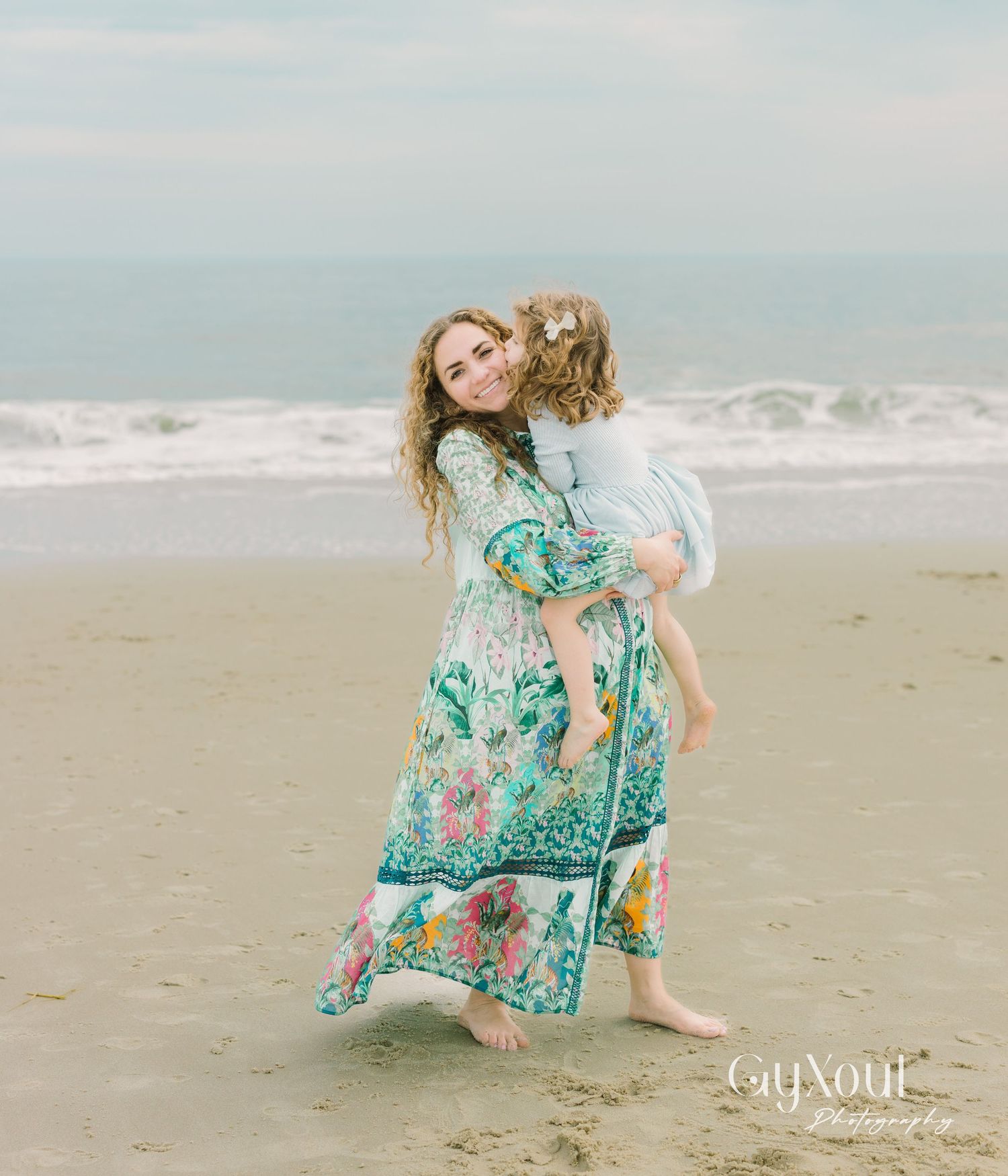 A pair in flowing turquoise floral dresses dance joyfully together on a misty beach shoreline.