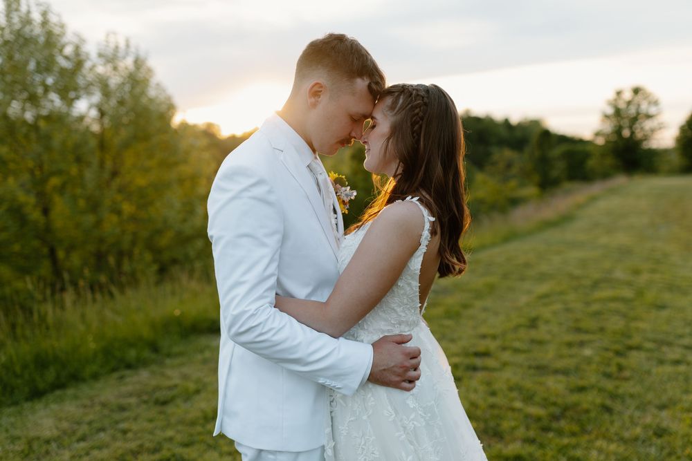 A couple in wedding attire shares a romantic moment at sunset in a lush green field.