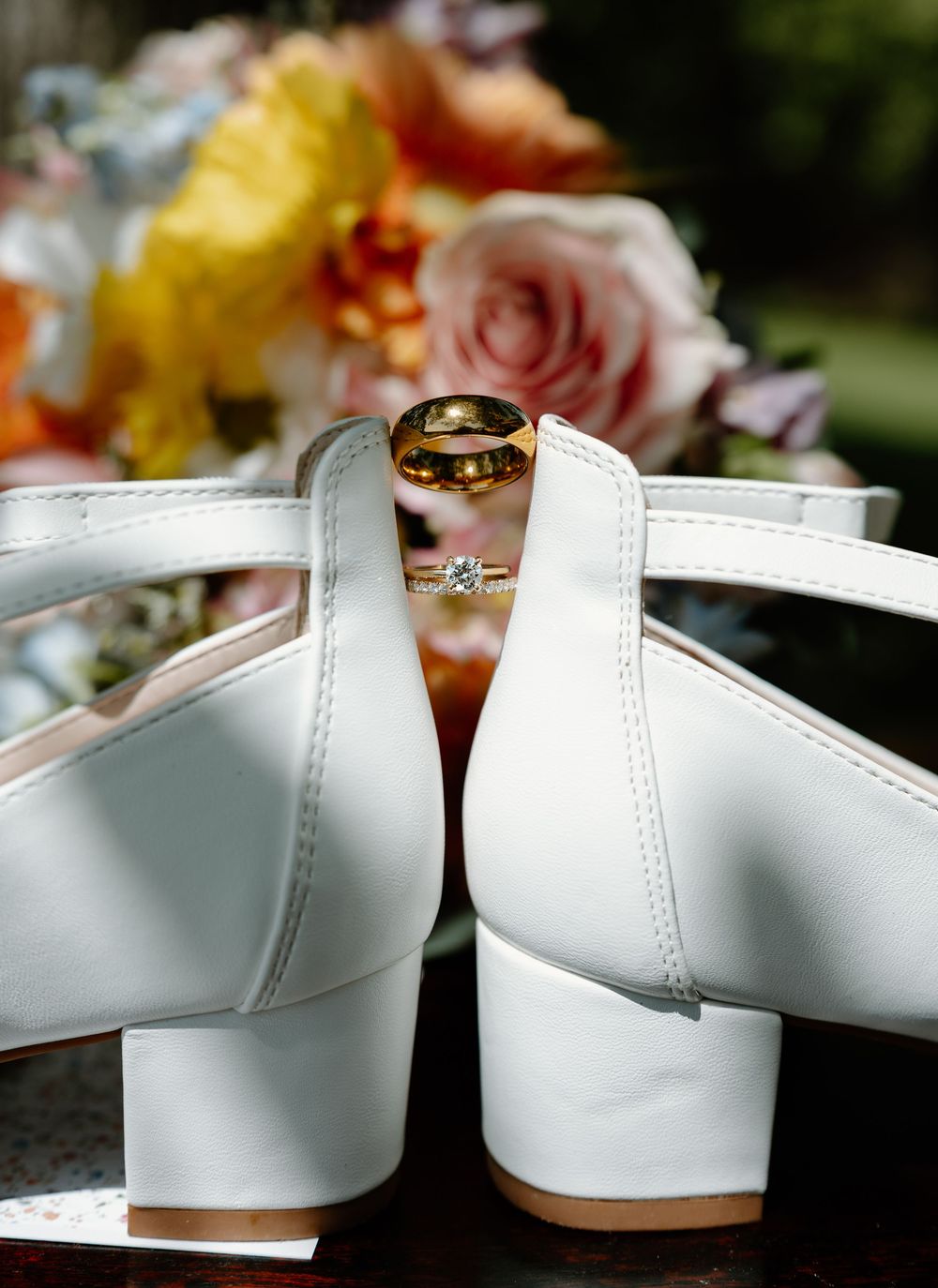 White bridal heels with gold wedding bands and diamond engagement ring, displayed against colorful floral bouquet backdrop.