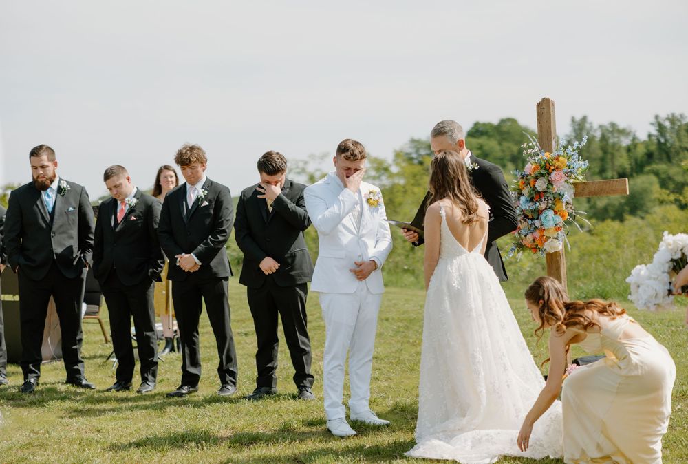 Outdoor wedding ceremony with groom in white suit, bride in white gown, groomsmen in black suits bowing heads in prayer by floral cross.