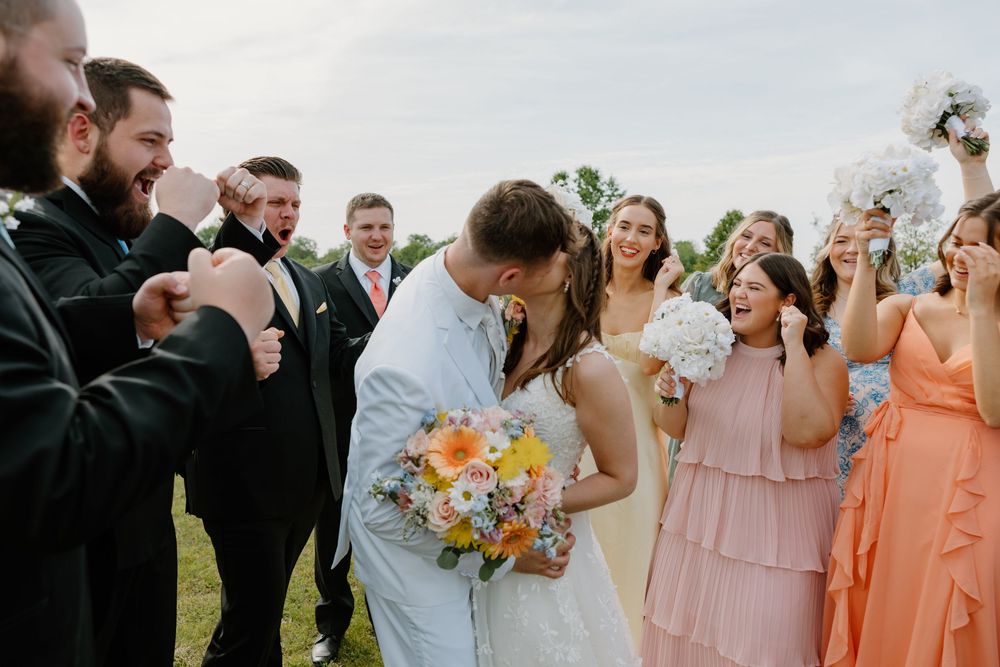 Bride and groom share a kiss surrounded by cheering wedding party in pink bridesmaid dresses and black tuxedos outdoors.