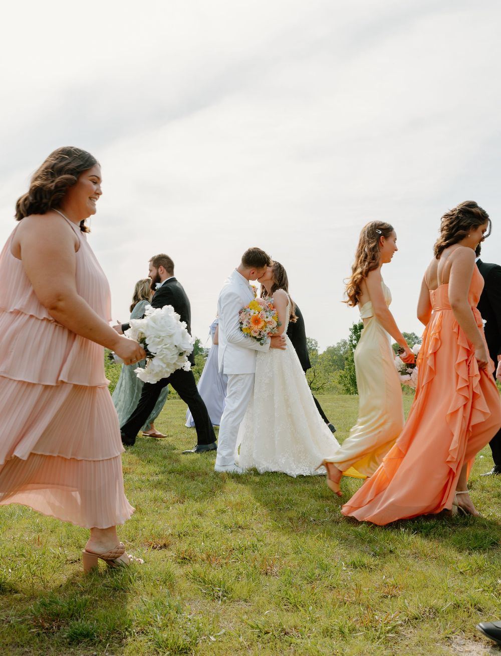 Bride and groom share a kiss surrounded by bridesmaids in peach gowns during an outdoor wedding ceremony.
