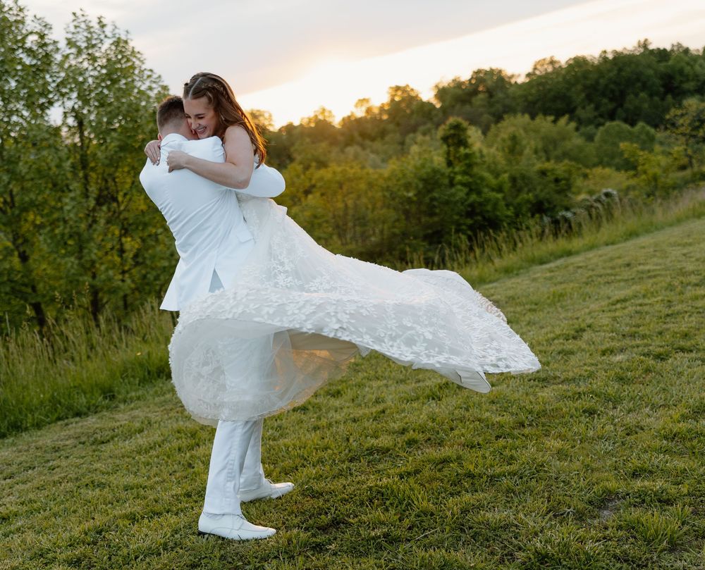 Groom lifting bride in white lace wedding dress at sunset on green hillside, romantic outdoor wedding photo.
