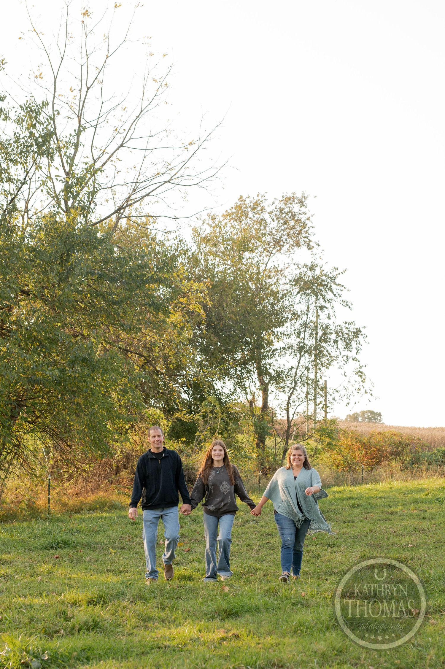 Three people hold hands while walking through a grassy field with autumn trees.