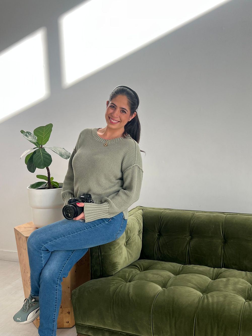 Female photographer sitting on green velvet sofa holding camera, smiling in bright natural light.
