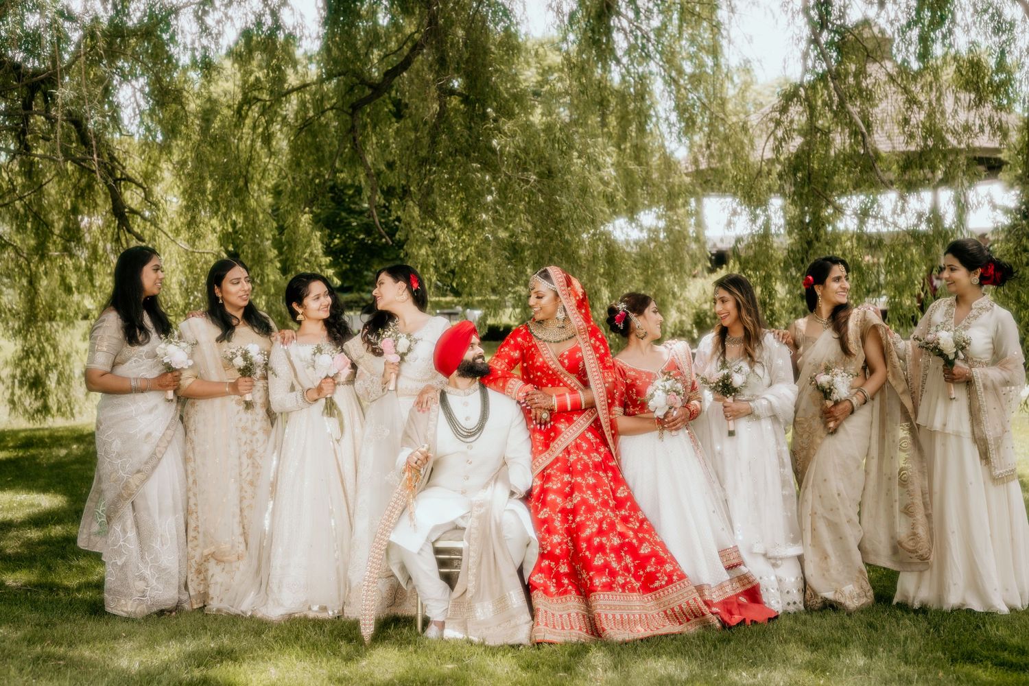 A lively wedding party poses on grass under willow trees wearing white dresses with one person in vibrant red.