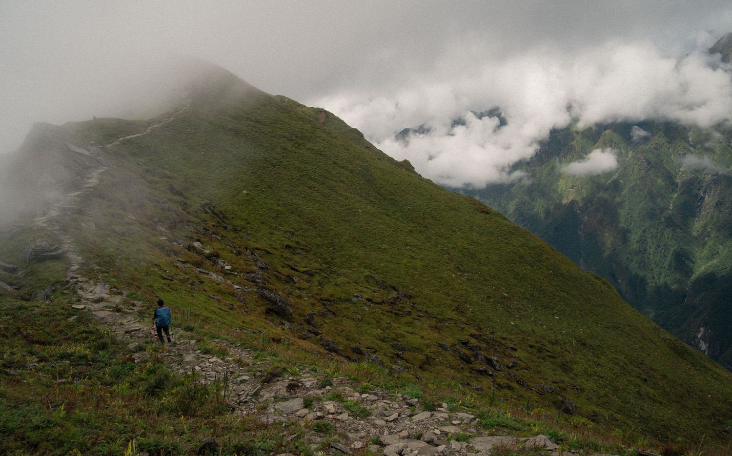 Scenic mountain trail winding through green slopes with dramatic cloudy peaks in the distance.