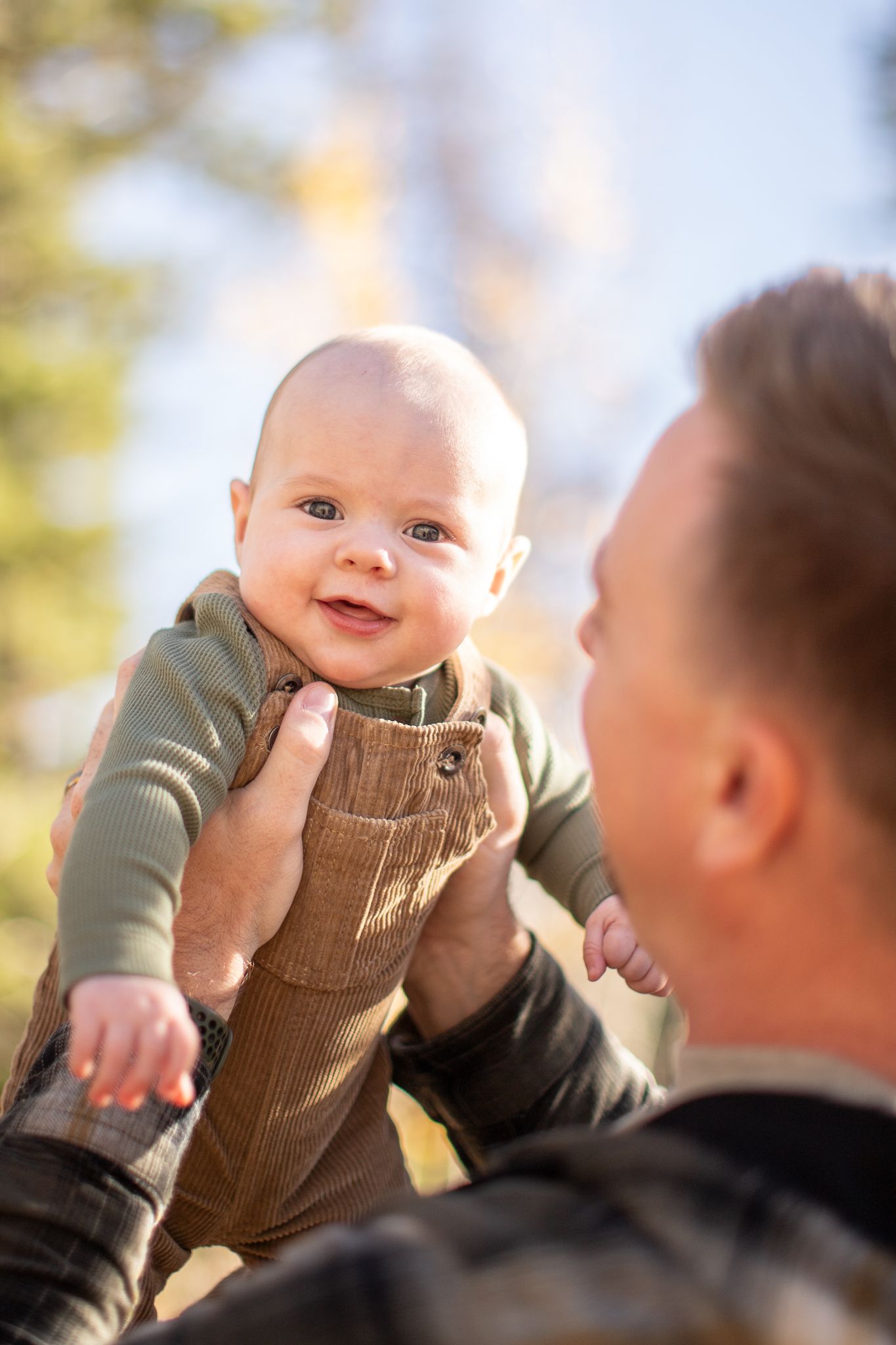 A happy baby wearing brown corduroy overalls smiles while being lifted up outdoors during a sunny day.