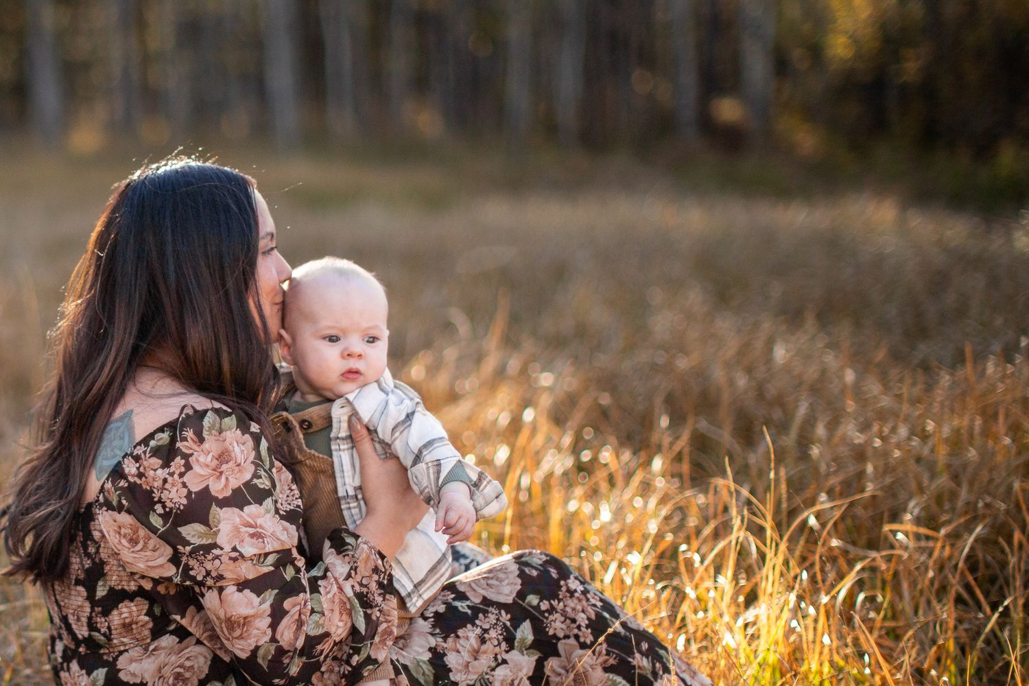 Parent and baby sit together in a golden autumn field at sunset as sunlight illuminates the tall grass around them.