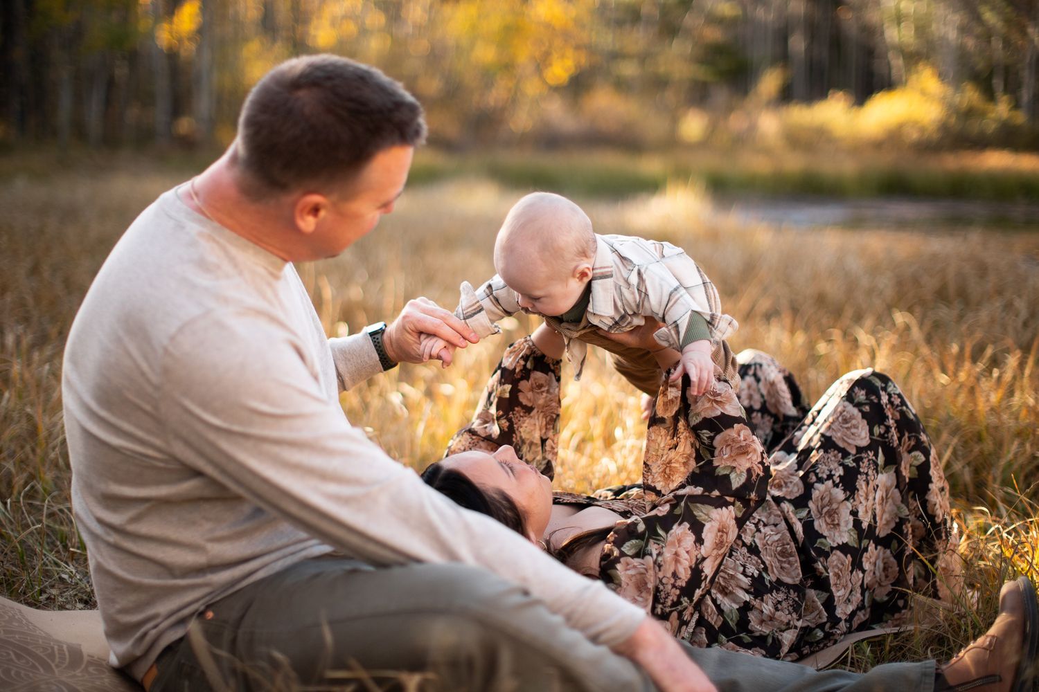A tender outdoor moment between a family during golden hour in a grassy meadow with autumn foliage in background.