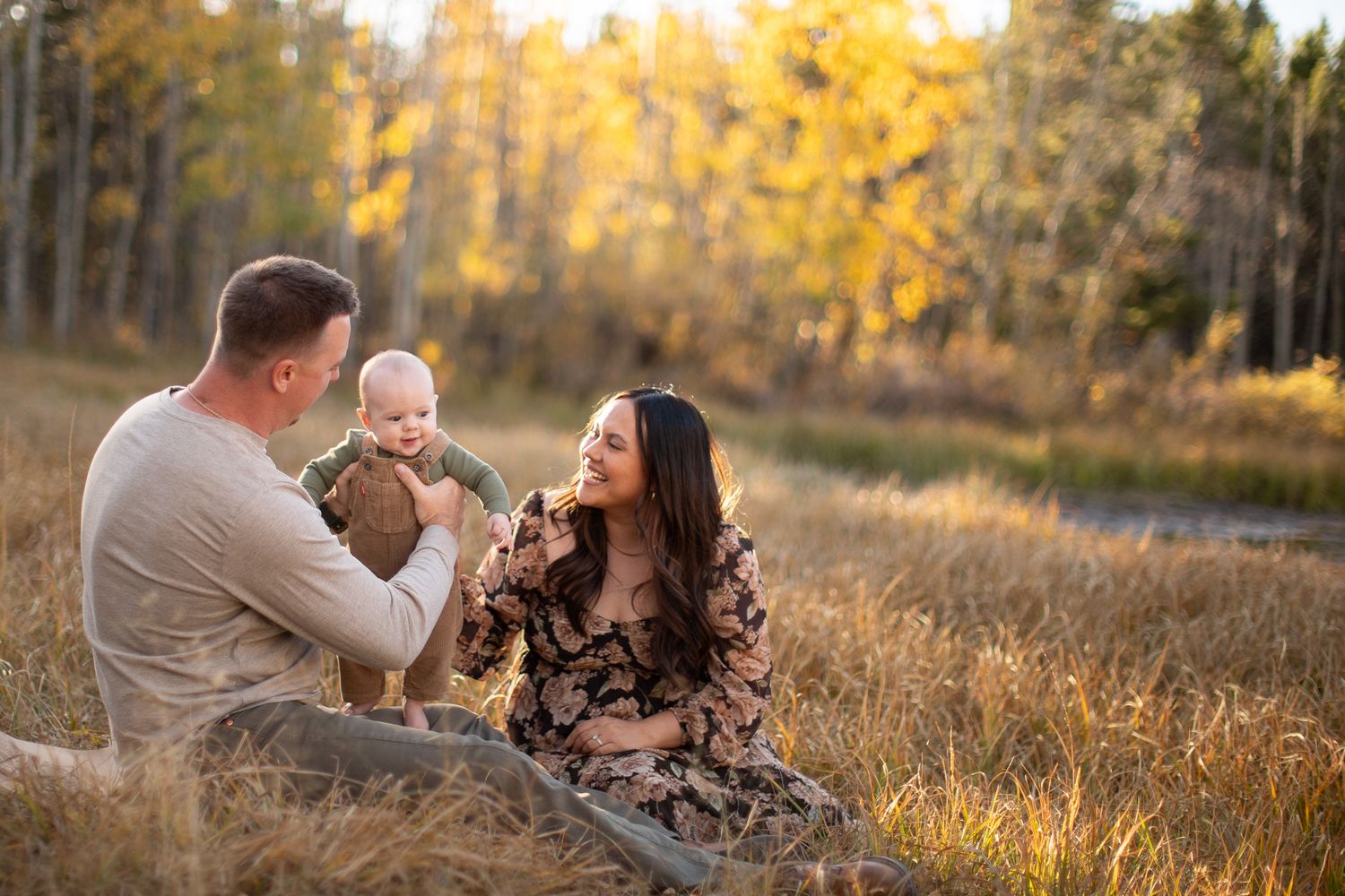 A family shares a tender moment together while sitting in a sunlit meadow with golden autumn trees in the background.