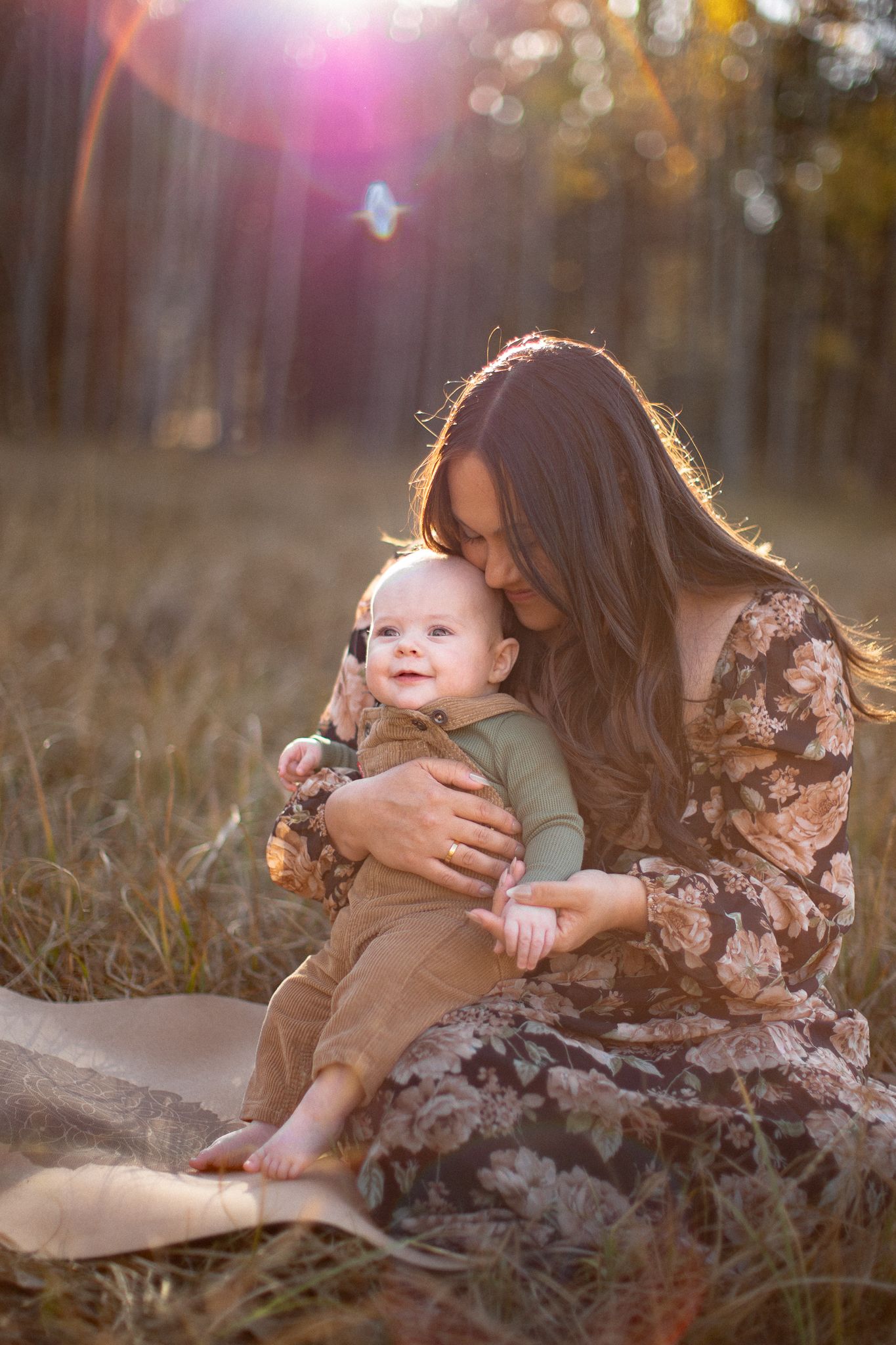 An outdoor portrait of a loving embrace between mother and baby on a blanket during golden hour in nature with sunflare.
