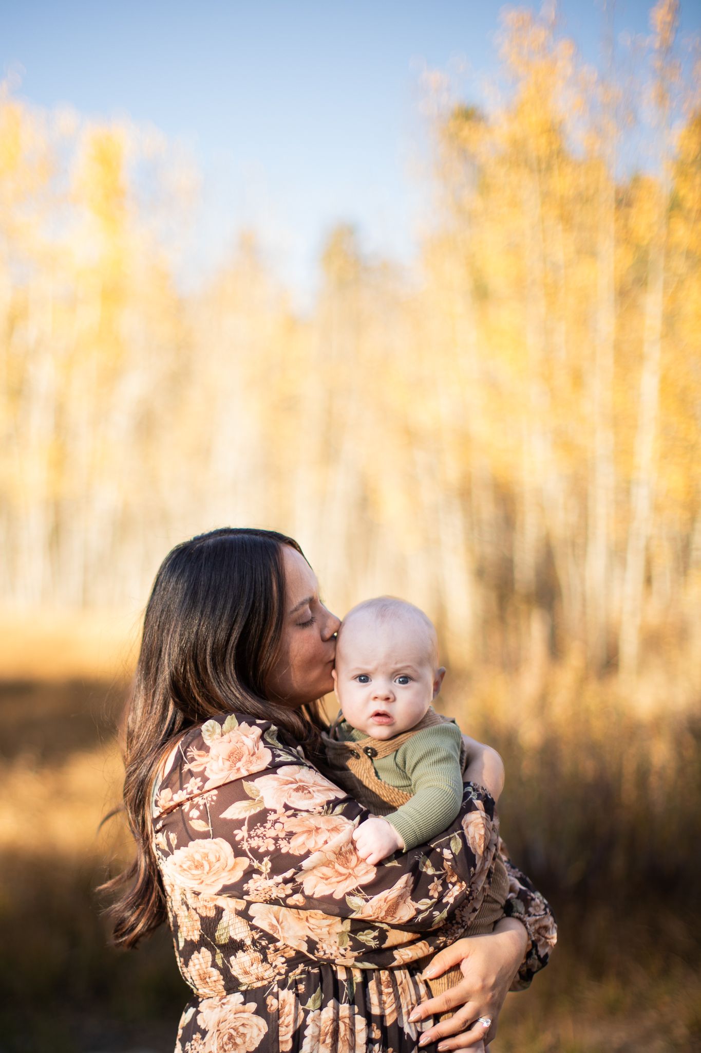 A parent holds and kisses their baby against a backdrop of golden sunlit trees during golden hour.