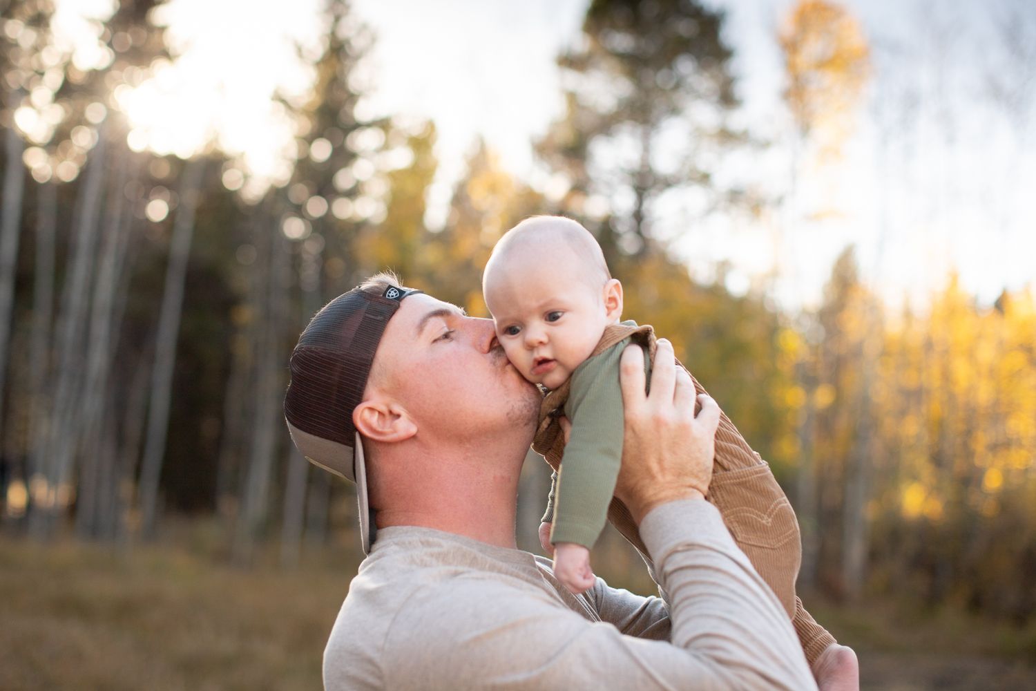 A parent in a beanie holds up a baby against golden autumn sunlight filtering through trees in a forest setting.