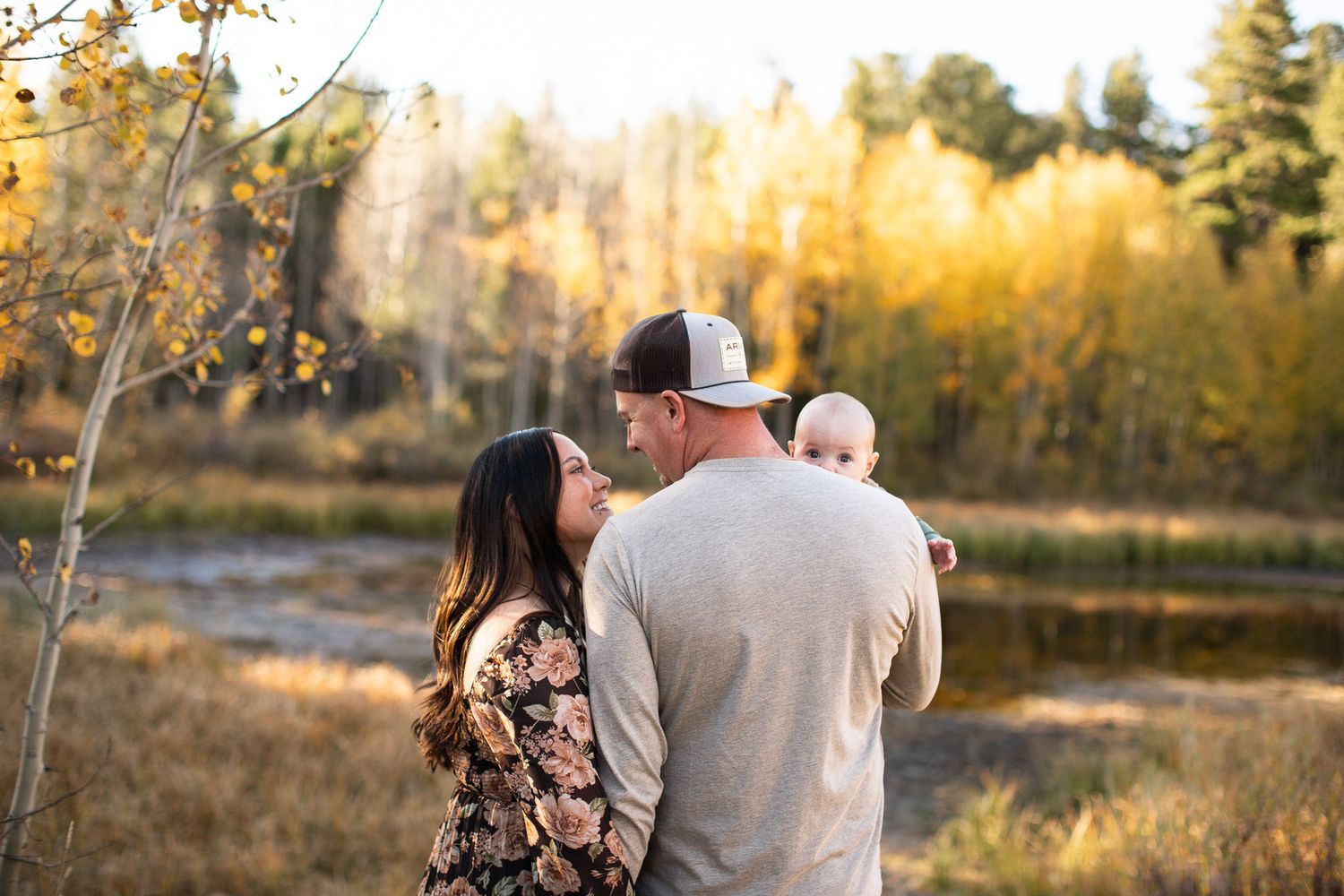 A family stands together outdoors during golden hour, surrounded by autumn foliage near a tranquil pond.
