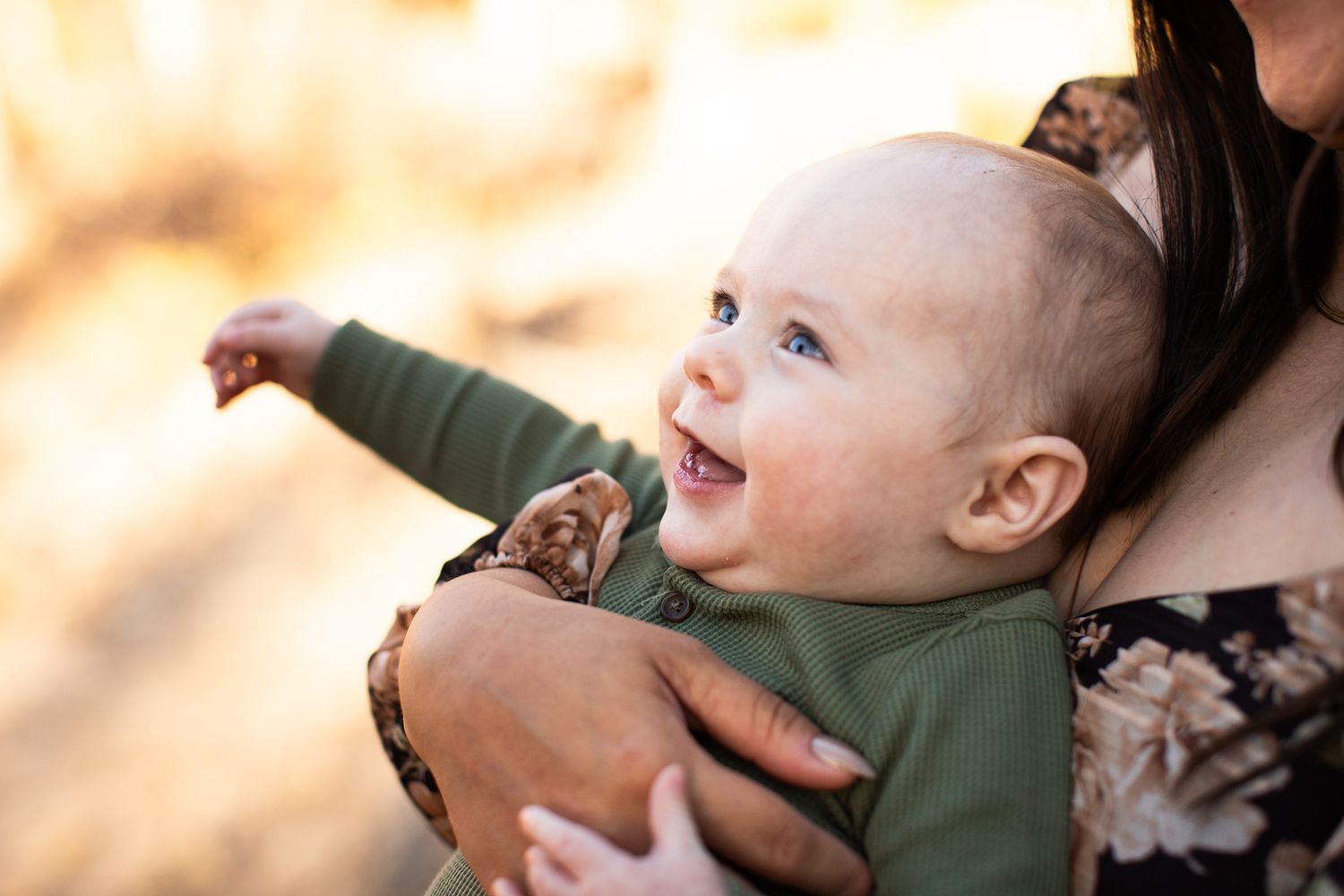 Happy laughing baby wearing a green outfit being held outdoors during golden hour lighting.