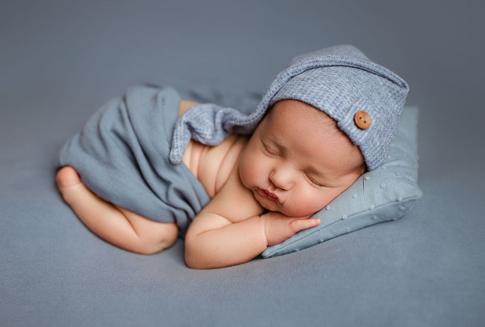 Newborn baby sleeping peacefully in blue wrap and knit hat on gray background.