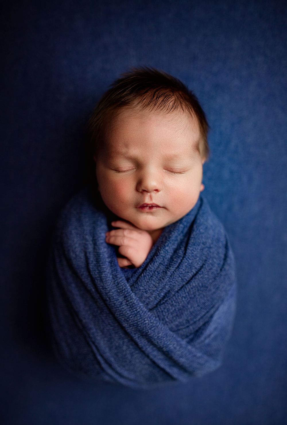 Sleeping newborn baby swaddled in a blue wrap against a dark blue background in a professional portrait.