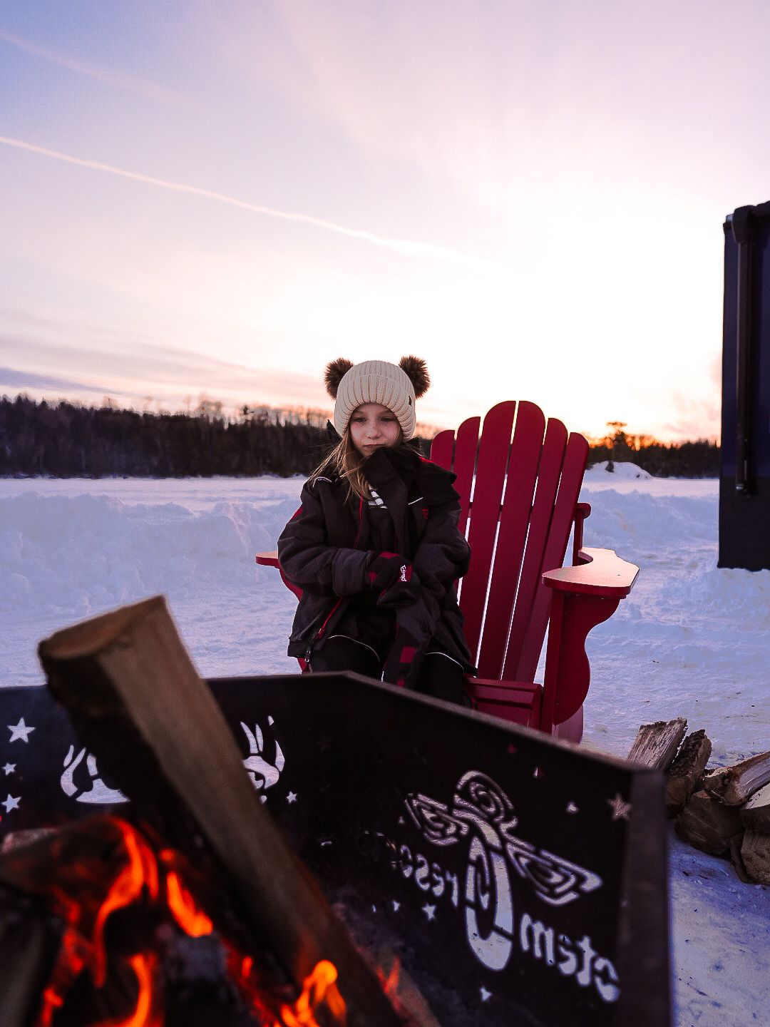 Winter scene with red Adirondack chair and campfire against snowy landscape at sunset.