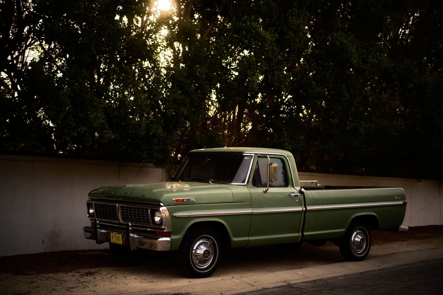 Classic green Ford F-100 pickup truck parked at dusk with sunlight filtering through trees in background.