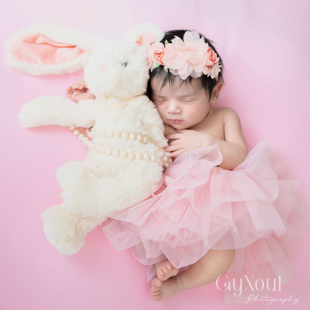 Sleeping newborn in pink tulle dress cuddling white teddy bear on pink background.