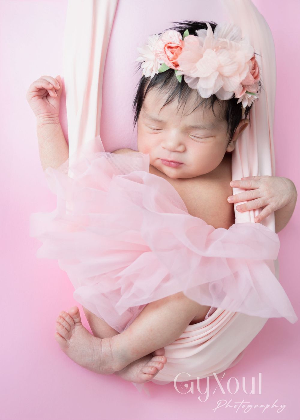 Peaceful newborn wearing floral headpiece and pink tulle dress against pink backdrop.