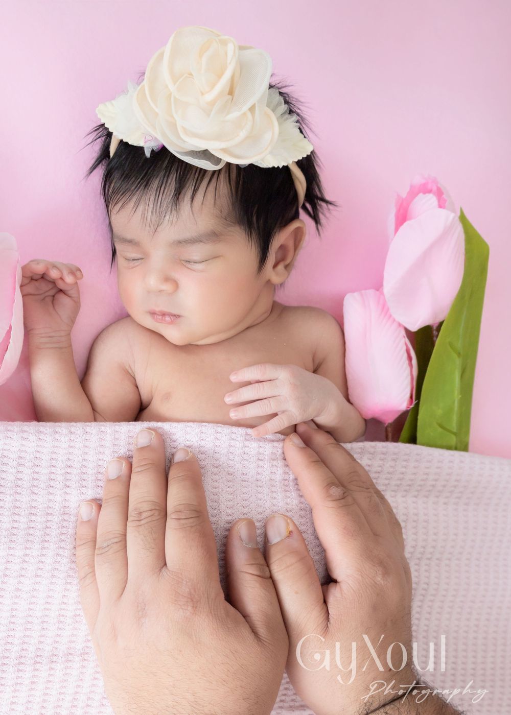 Sleeping newborn with white rose headpiece beside pink tulips on textured blanket.