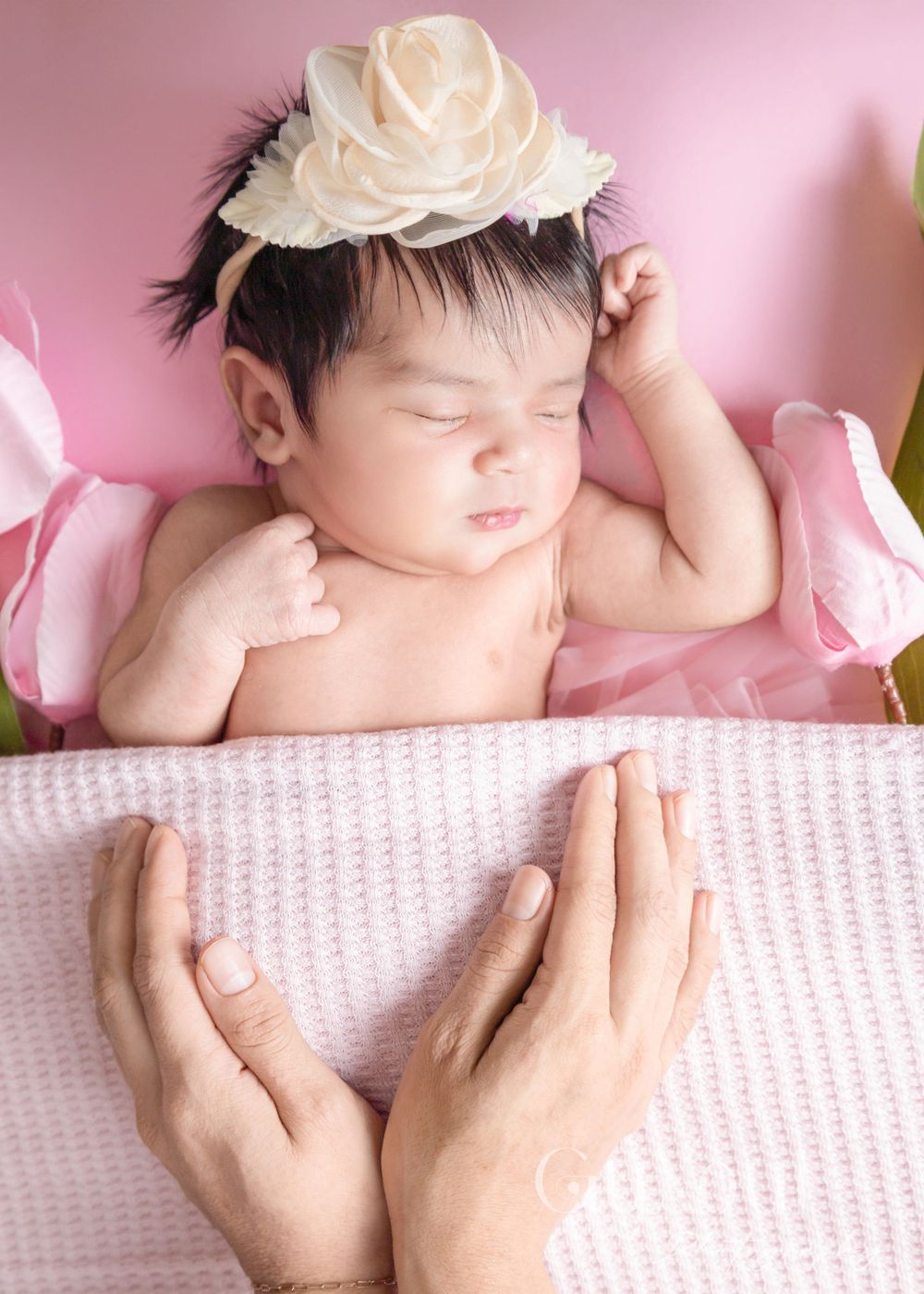 Gentle hands supporting sleeping newborn wearing cream rose headpiece with pink tulips.