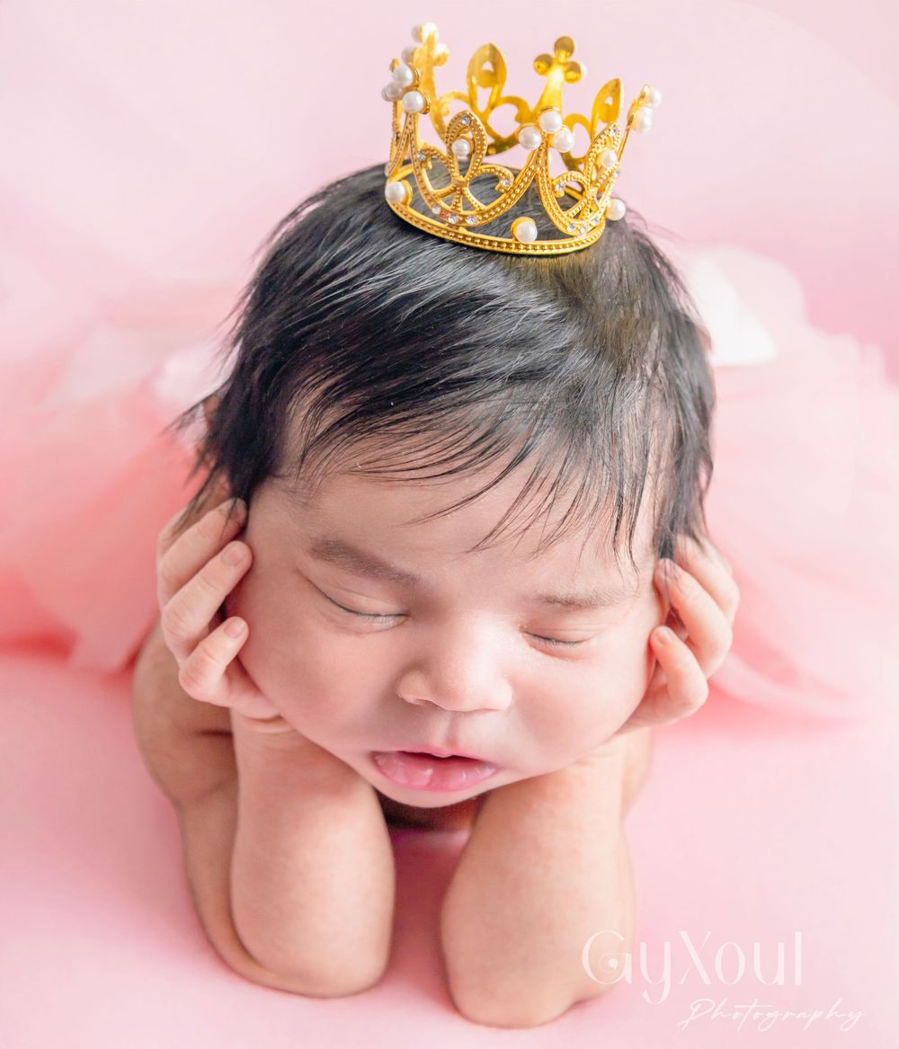 Sleeping newborn wearing golden crown against pink background.