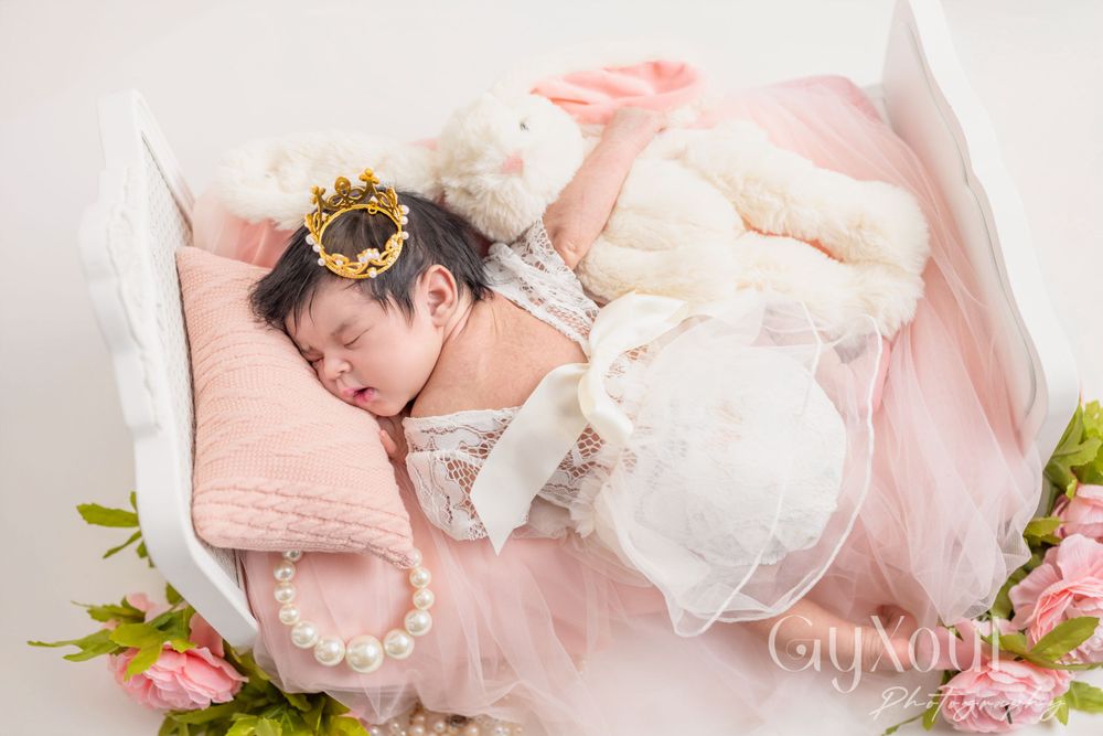 Newborn wearing a crown rests peacefully on pink bedding with floral accents in a dreamy photography setup.