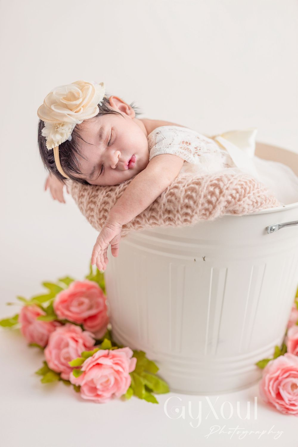 Sleeping baby posed in white bucket with pink roses and knit blanket in soft natural light.