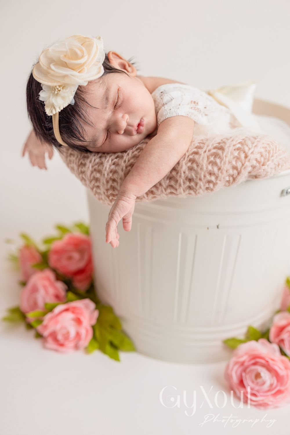 Peaceful newborn sleeping on white bucket surrounded by pink roses in delicate portrait setup.