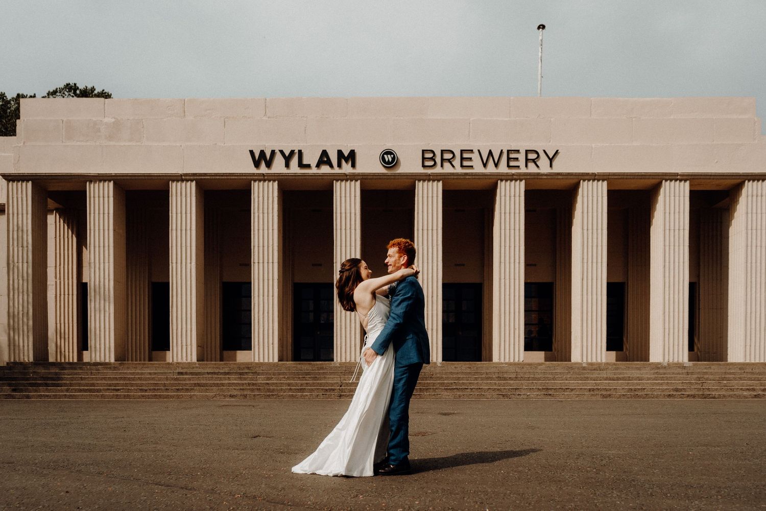 Two people in wedding attire embrace in front of a columned building marked 'Wylam Brewery' at golden hour.