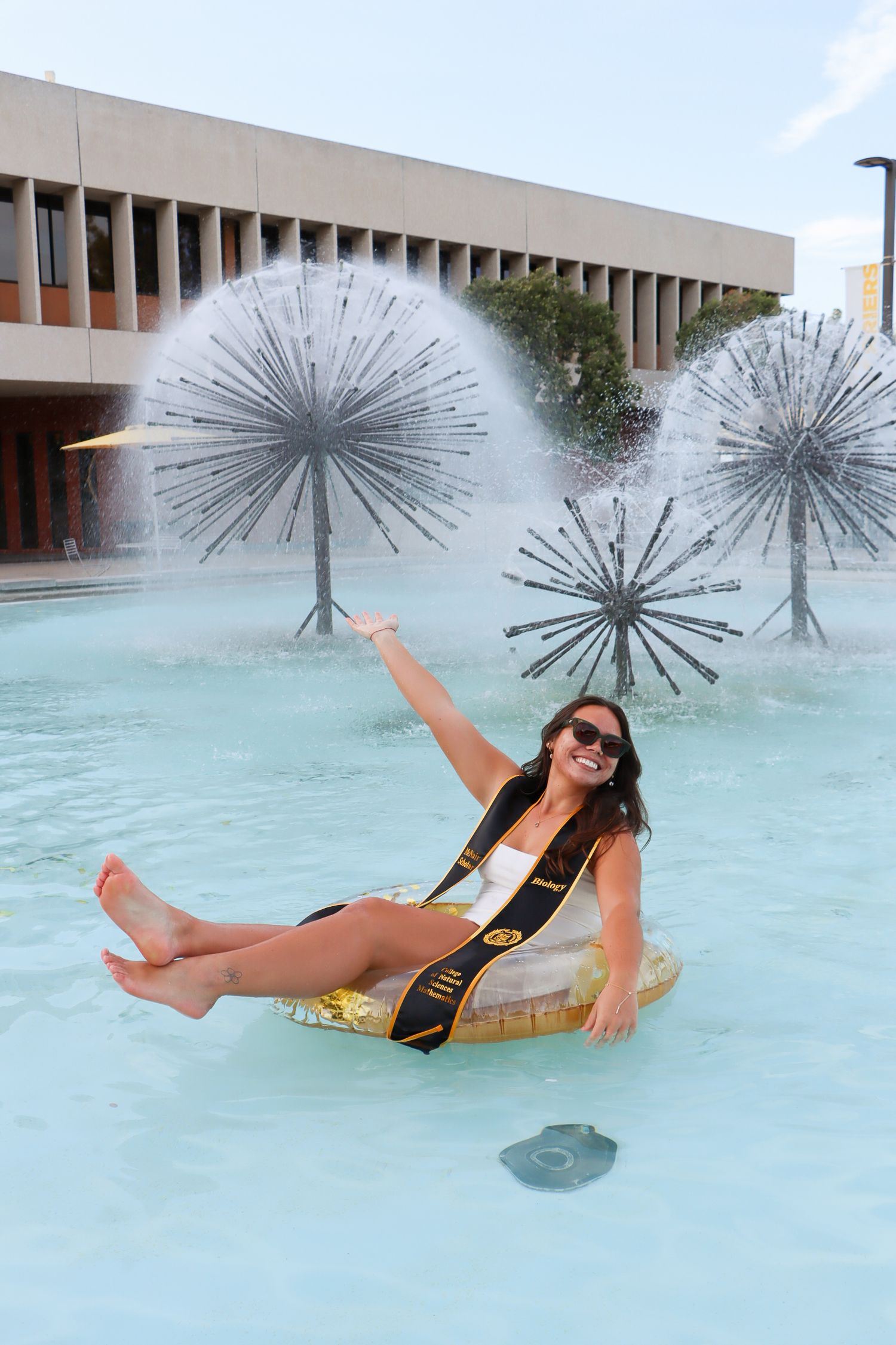 Graduate lounging in campus fountain with decorative metal sculptures while wearing academic regalia.
