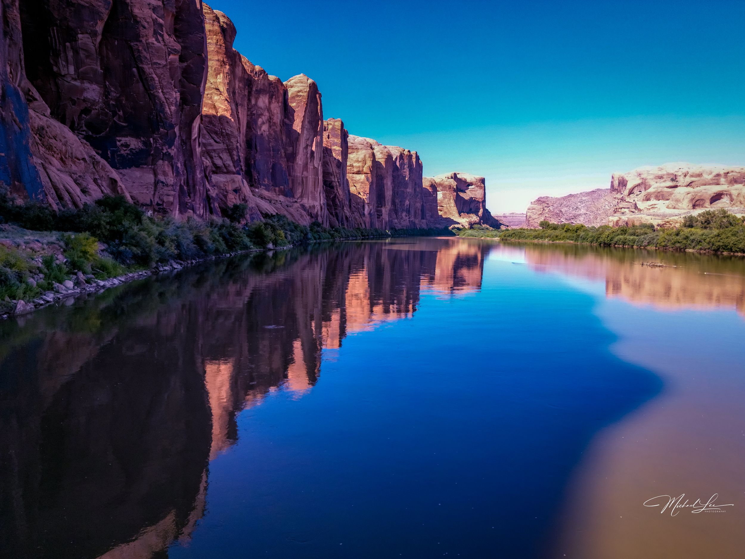 Red rock cliffs perfectly reflected in calm river waters under blue sky.