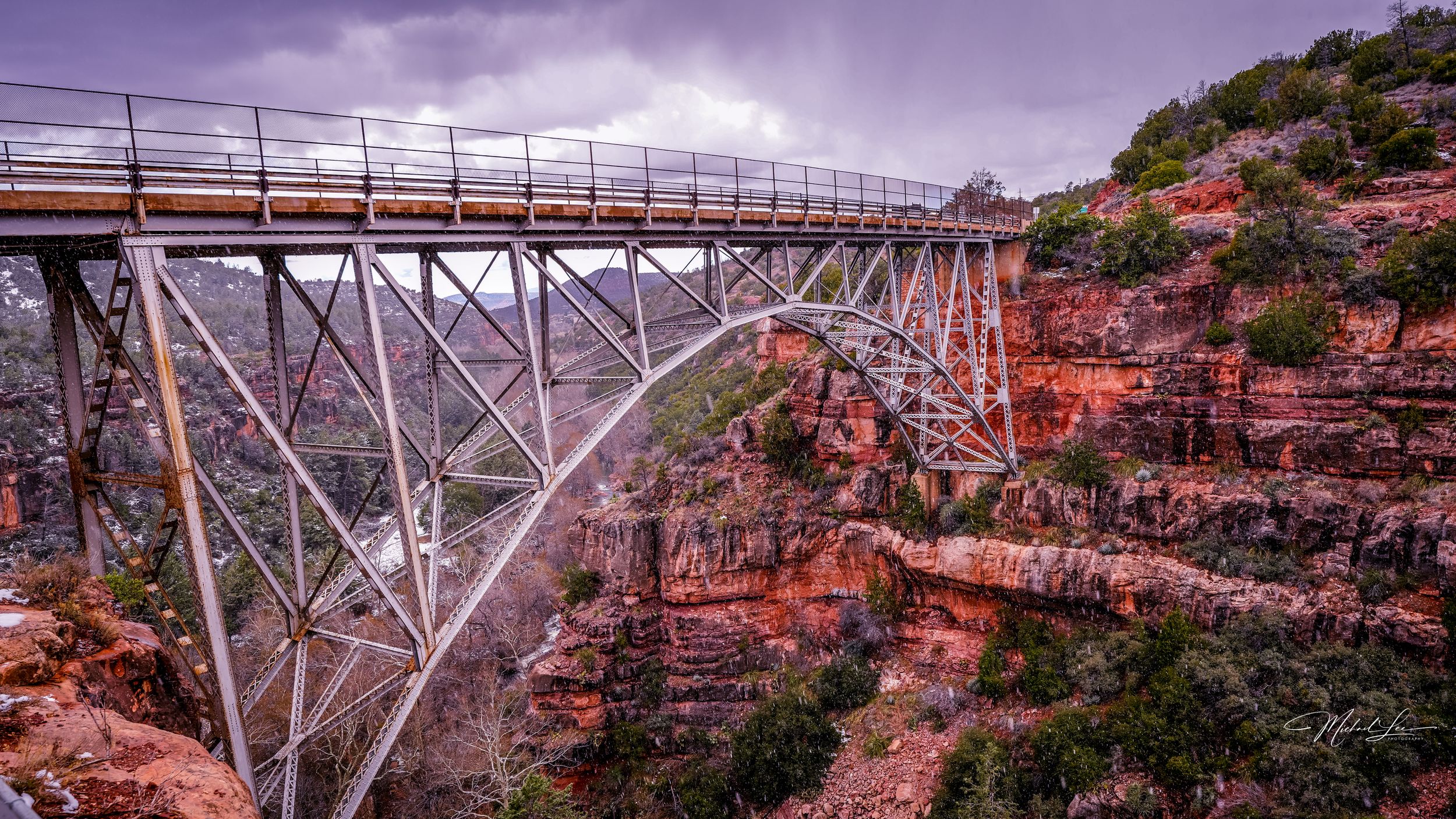 Steel arch bridge spanning across red rock canyon under dramatic purple sky.