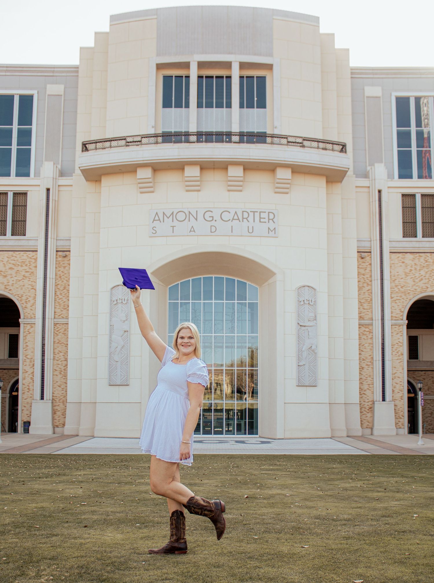 A graduate wearing a light blue dress and boots poses while holding up a diploma in front of the Amon Carter building.
