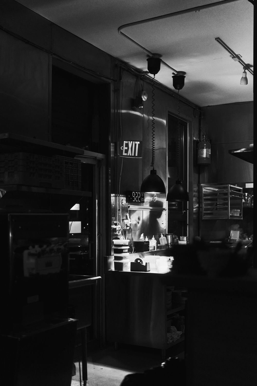 Dark restaurant kitchen interior with exit sign illuminated and industrial lighting fixtures.